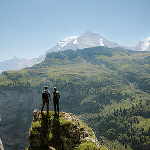 Zwei Wanderer stehen auf einem Felsvorsprung und betrachten die grüne Berglandschaft mit imposanten Gipfeln.